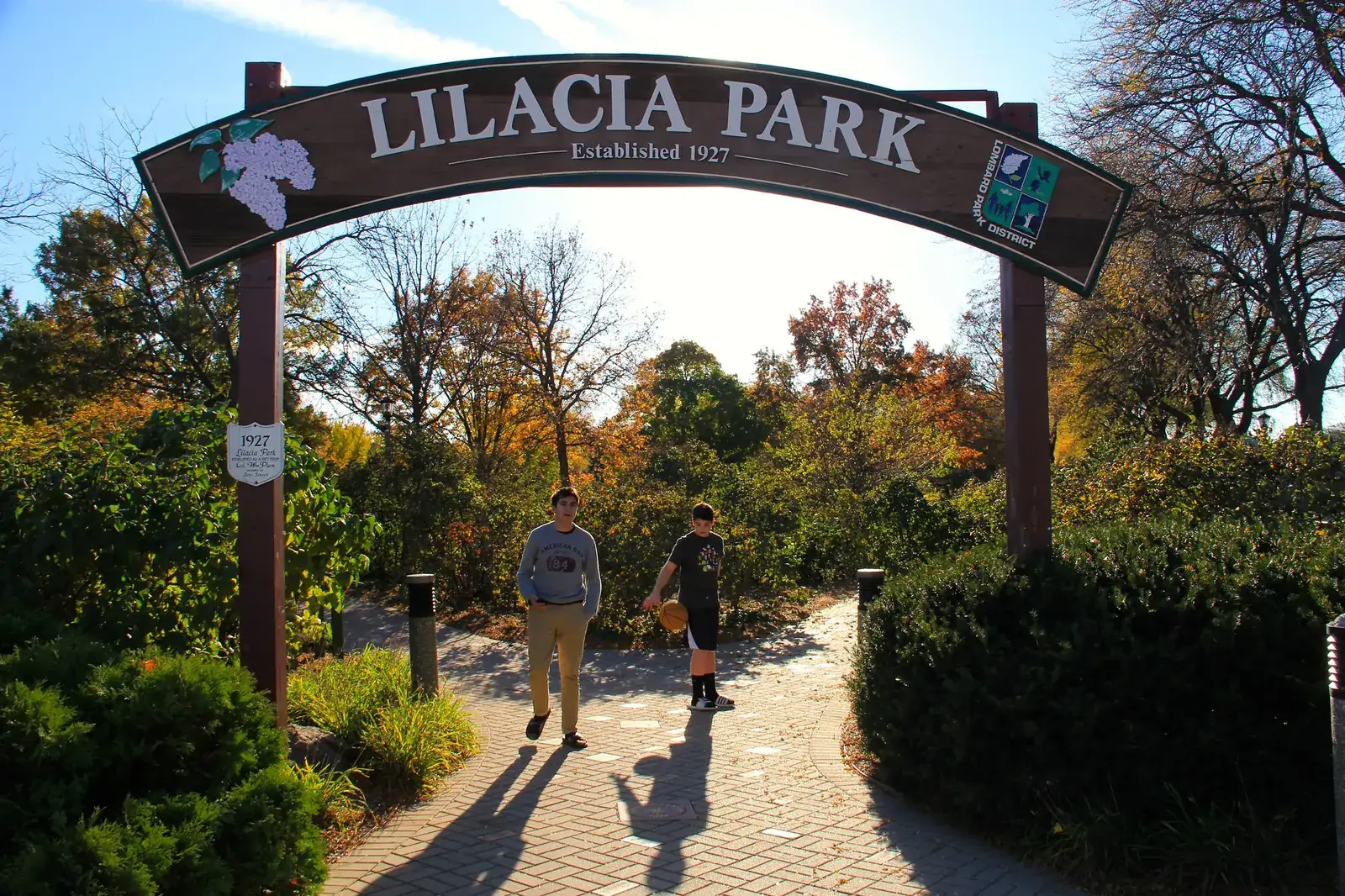 The arched Lilacia Park sign over a brick path, with trees in fall color and two people walking in.