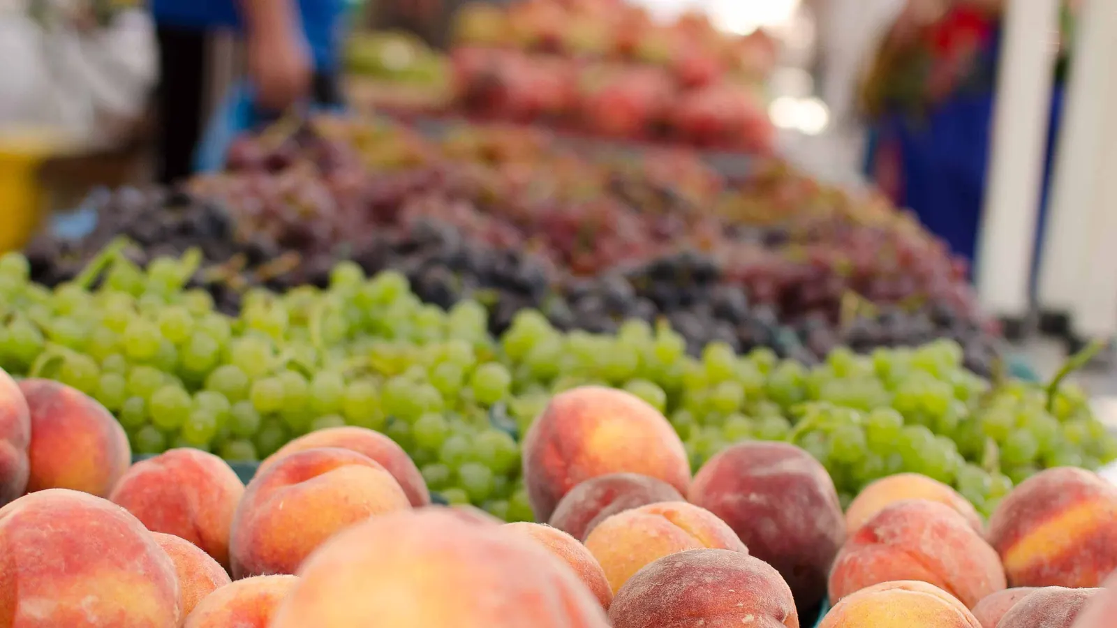 Peaches and green and red grapes stacked on a farmers market table, with shoppers behind.