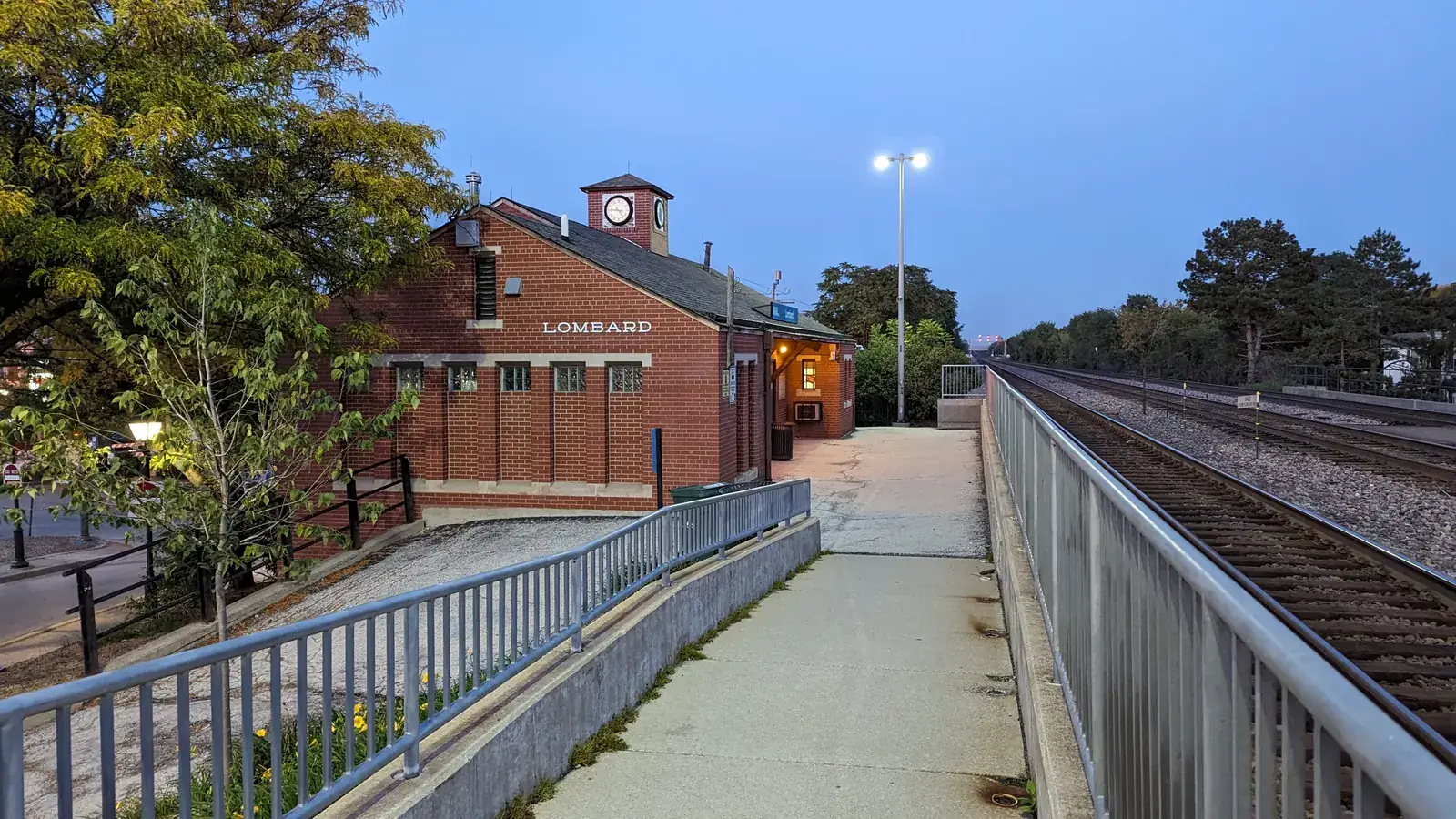 The red-brick Lombard Metra station with its clock tower, beside the platform and tracks at dusk.