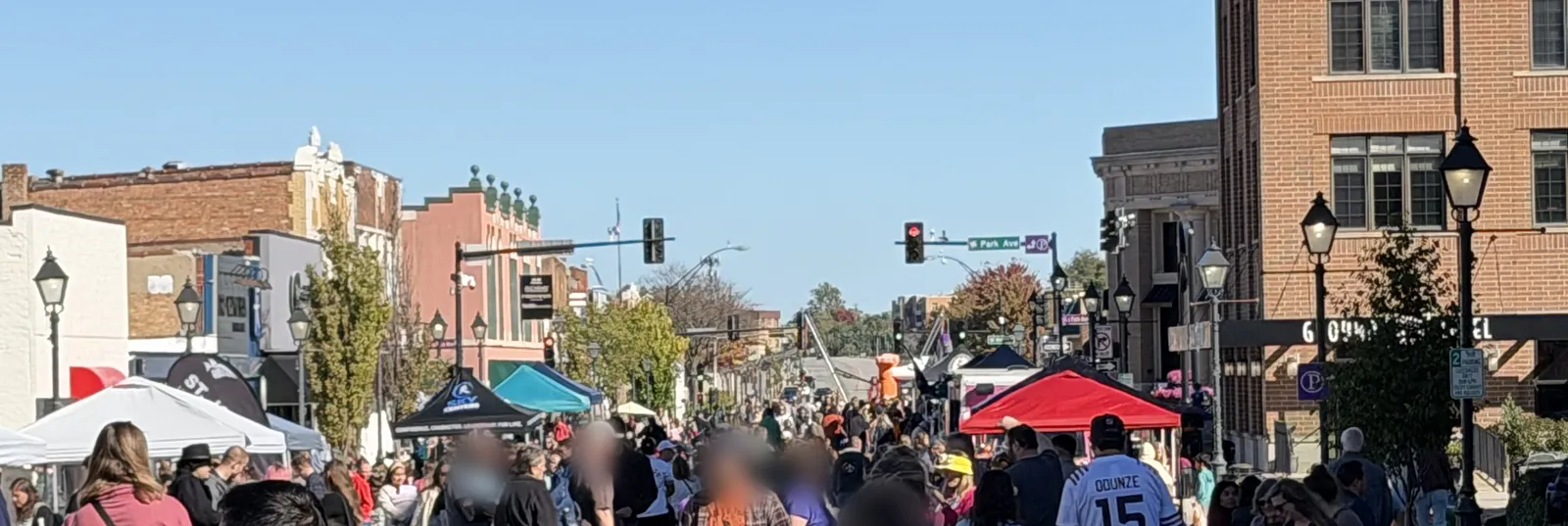 A crowd fills a closed downtown Lombard street between brick storefronts under a clear blue sky, with vendor tents including a red canopy and a teal canopy, old-fashioned street lamps, and the Park Avenue street sign in the distance.